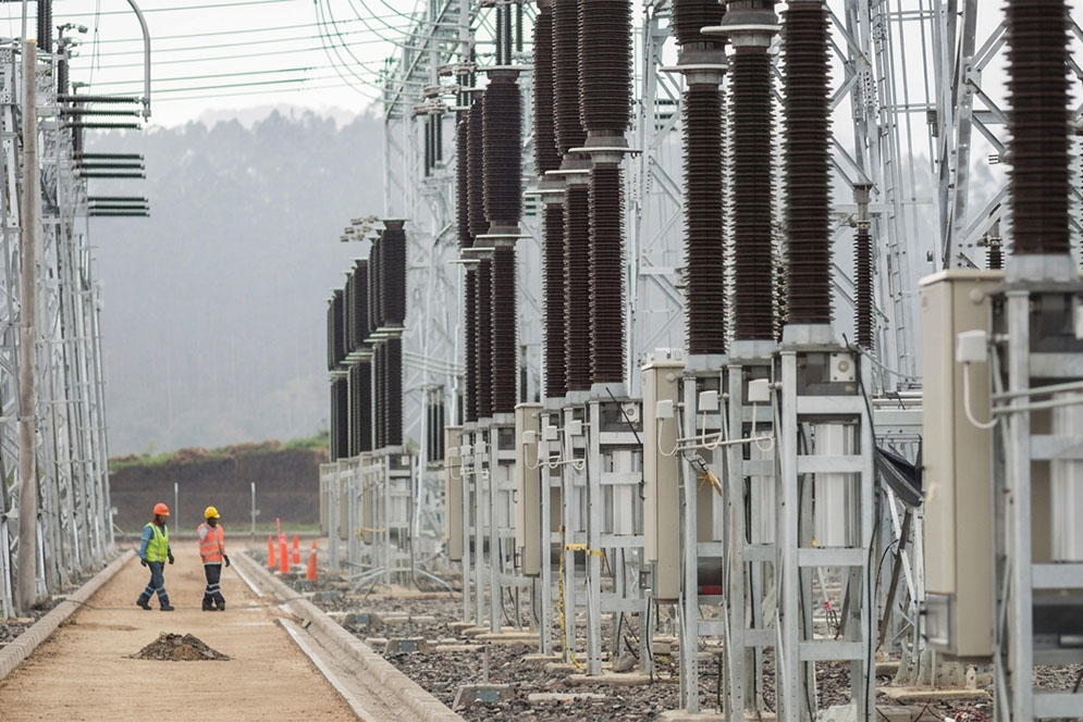 Dos operarios recorren un patio de subestación eléctrica con hileras de aisladores de alta tensión.