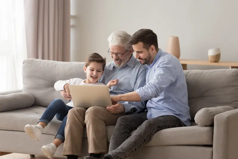 Padre, hijo y nieto sentados en un sofá, sonriendo frente a un computador, al revisar las características del plan de Asistencia Funeraria de Enel.