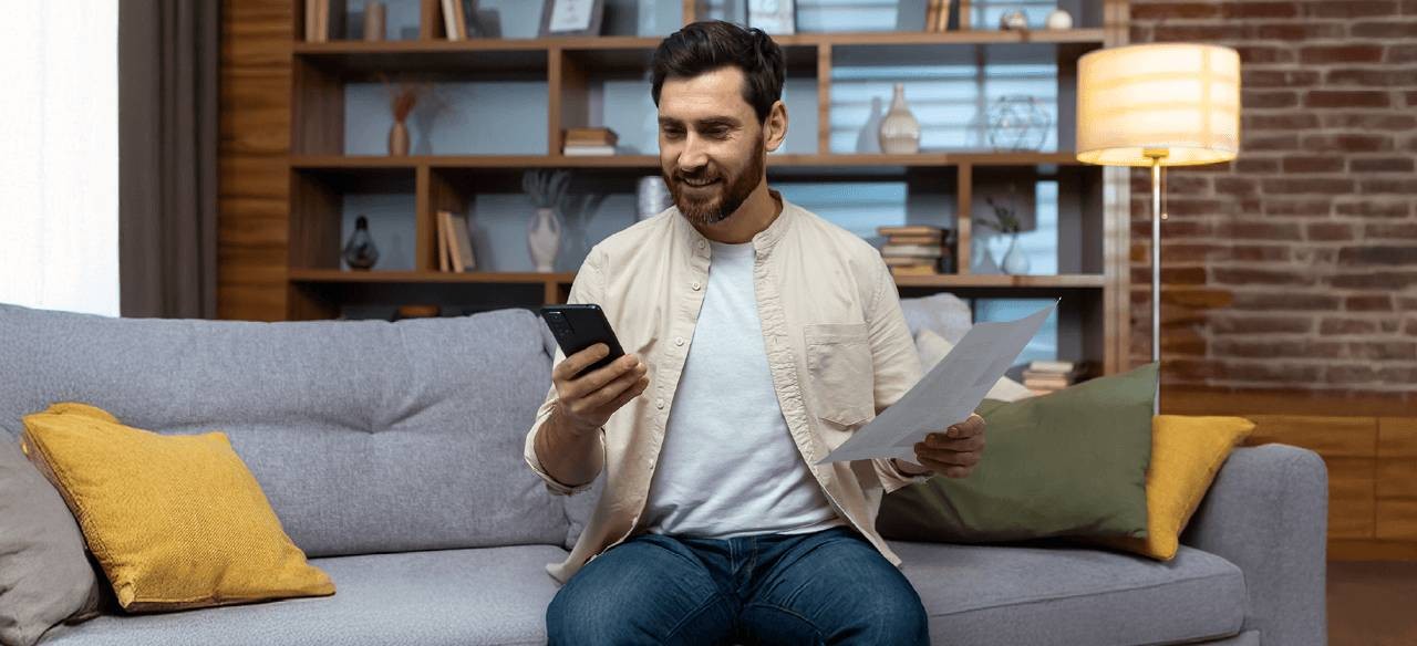 Hombre revisando su celular mientras sostiene una factura de energía en su sala.