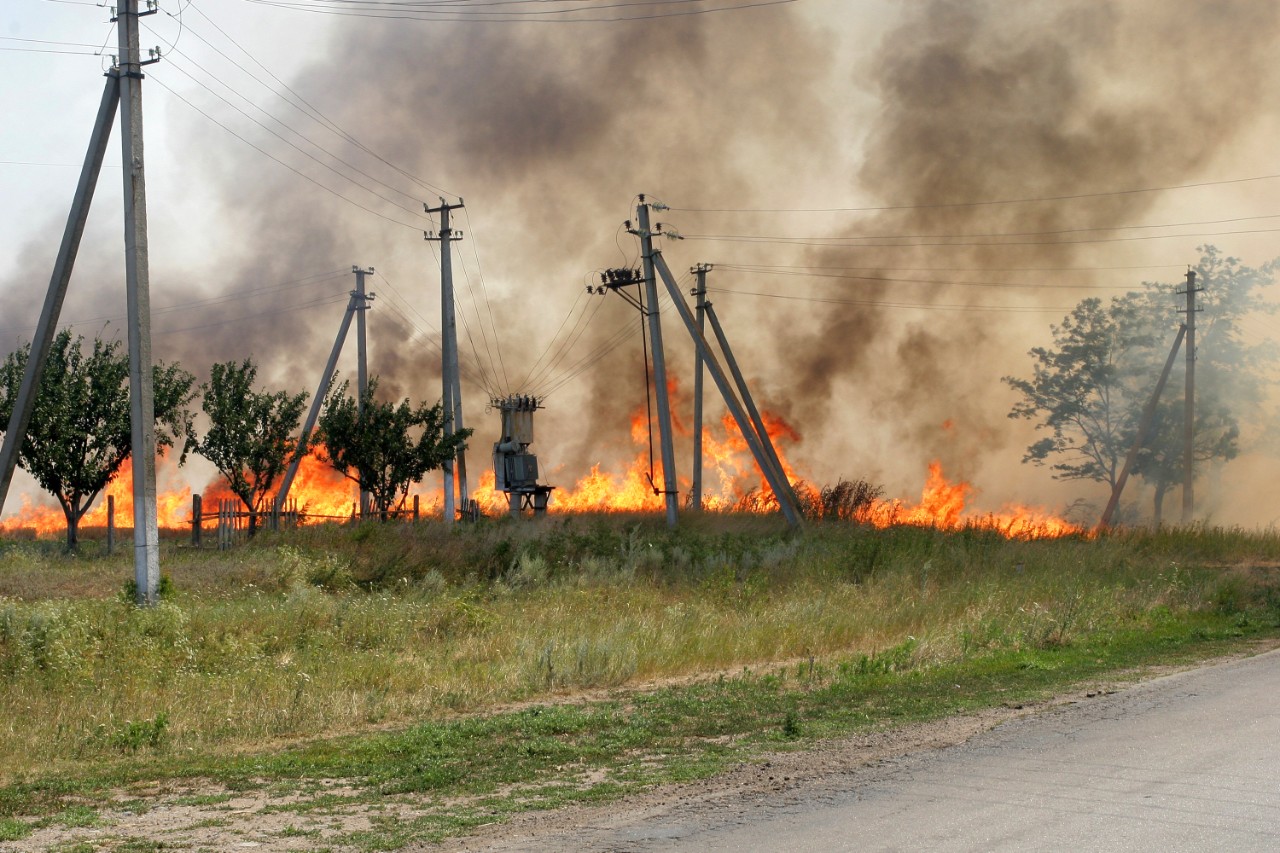 Incendio de vegetación seca junto a una carretera rural avanza al lado de postes y redes eléctricas.