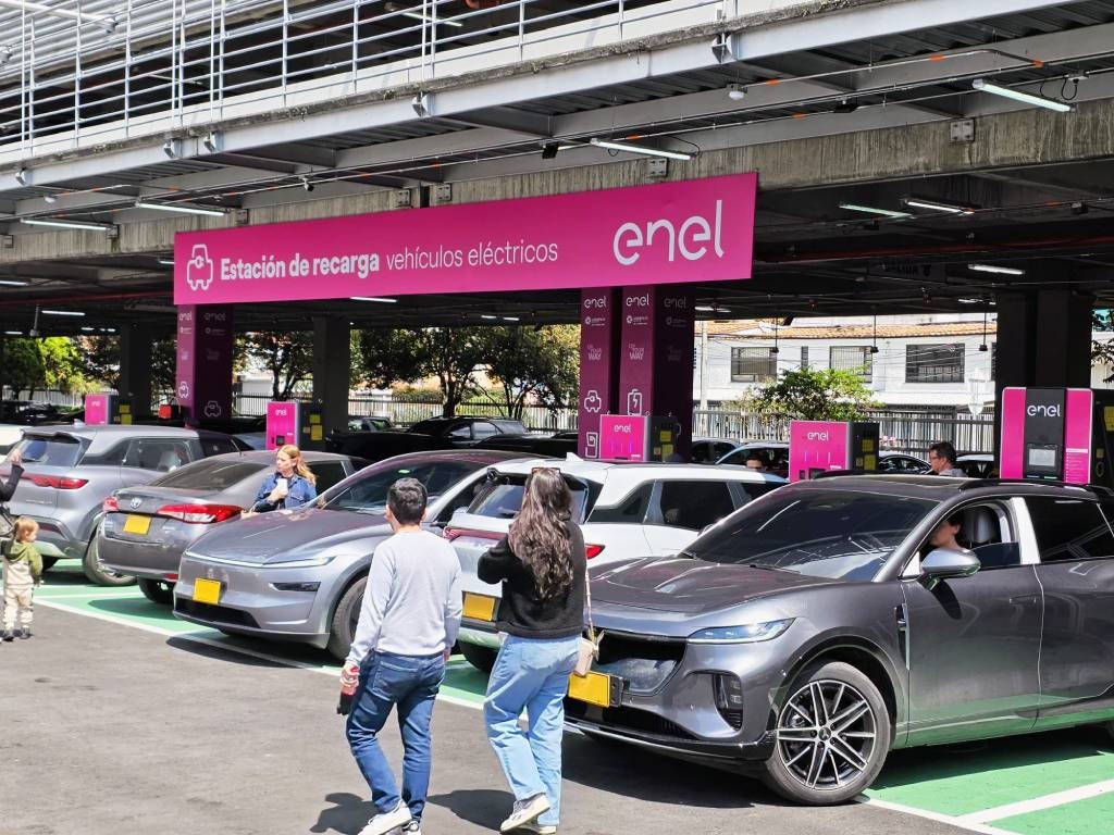 Personas caminan junto a carros eléctricos conectados en una estación de recarga de Enel en un parqueadero cubierto