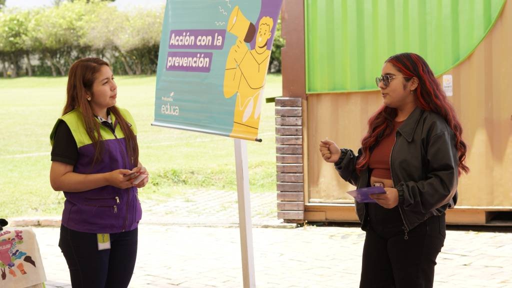 Dos mujeres conversan junto a un aviso de prevención en una actividad comunitaria al aire libre
